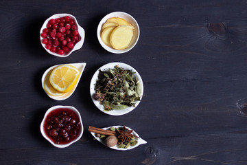 raspberry jam, fresh cranberries, slices of ginger and lemon, herbal tea leaves, spices in white plates as healthy organic treatment on black wooden background, top view with copy space