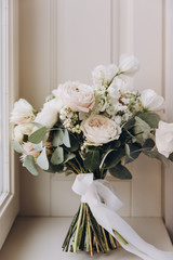 bride’s wedding bouquet of flowers and greenery with a ribbon stands near the window on a white...