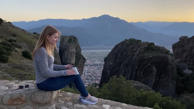 Young female tourist works remotely at laptop at Meteora mountains. Beautiful caucasian woman-freelancer works outdoor at computer in front of high rocks at sunset
