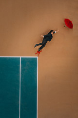man in a suit reaching for a red umbrella flying away while standing on a red chair on a tennis court
