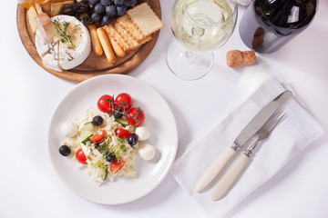 vegetables salad on the plate, glass of white wine and plate with assorted cheese, fruit and other snacks.