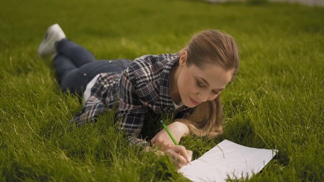 attractive nice girl lies on the grass and takes notes in a notebook. chewing gadget looks at the camera and smiles. 4K