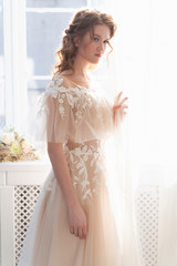 Beautiful natural redhead girl bride, with nude makeup, wearing a white dress, stands at the window next to a transparent curtain.