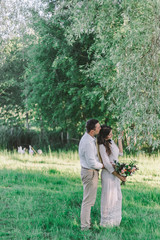 Wedding. The groom in a white shirt and light pants, the bride in a beautiful dress and a straw hat with a beautiful wedding bouquet in her hands. Hug each other look at a tree.