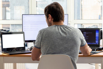 A man in a gray t-shirt is sitting at a workplace with two laptops and a monitor near the window....