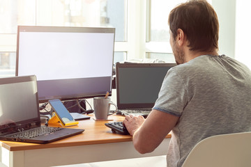 A man in a gray t-shirt is sitting at a workplace with two laptops and a monitor near the window....