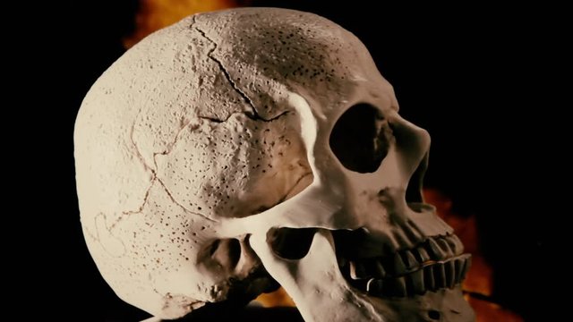 Front closeup shot of a scary human skull, rotating on its axis, over a fire pyre with a smooth crackling trail at night.