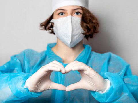 Medical Worker In A Disposable Mask And A Lab Coat Makes A Heart Sign. Gray Homogeneous Background. Conceptual Photo: Doctors On The Protection Of Health.