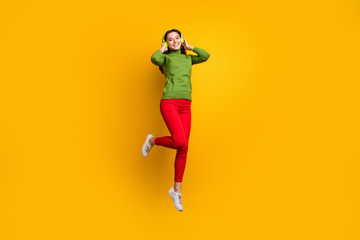 Fototapeta na wymiar Full size photo of funky pretty lady jumping high holding ears with cool modern headphones listening music wear casual green jumper red pants isolated yellow color background