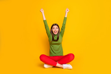 Photo of beautiful lady sit floor holding fists raised supporting favorite sports team excited cheery mood wear green sweater red trousers isolated yellow color background