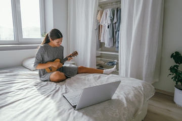 pretty young woman study to play ukulele guitar online at home  with notebook sitting ot the bed in...