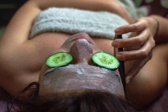Busy woman talking on the phone while using an anti aging face mask at home. Natural beauty treatment with cucumber and bio cream. - Powered by Adobe