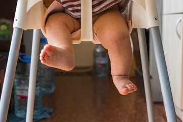 Cute legs hang from baby highchair at home. Infant sitting in chair.
