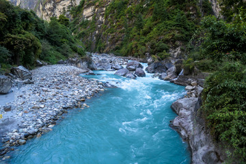 View on River and Mountains from Bhulbhule, Annapurna Circuit Trek, Nepal. Turquoise color of the river, big stones popping out of the river. Green forest around. Whole valley is shrouded in shadow.