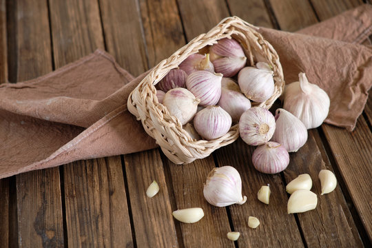 White round garlic falls from a wicker basket on a brown wooden background with a dark napkin. Close-up photo