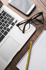 Espacio de trabajo o estudio en casa. Ordenador portátil, gafas de vista, teléfono móvil, lápiz y bloc de notas sobre mesa de madera.
