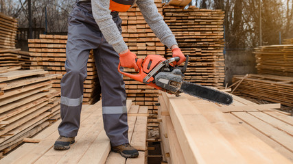 Fototapeta premium Close up young man carpenter at the wooden warehouse hold in his hands a chainsaw and saw wooden plank/board.