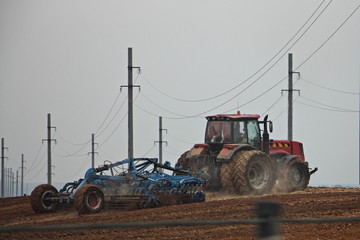 Red big twin wheels tractor with large plough ploughs the land in a field near the power lines...