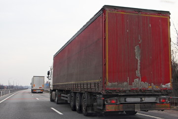 Old red semi truck move on two lane suburban asphalt highway at spring day, rear side view – international logistics, cargo transportation, trucking industry