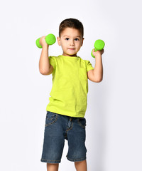 Little male in yellow t-shirt and denim shorts. He smiling, lifting green dumbbell, posing isolated on white studio background