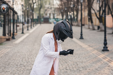thin girl in a white medical coat and overalls holds a syringe. A woman in a motorcycle helmet with a medical mask. On her hands are latex gloves. coronavirus, disease, infection, quarantine, covid-19