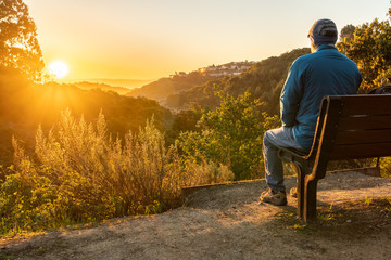 Person, man, sitting on a bench watching the sunrise over hills and valley with trees and shrubs in the middle and houses on a hill in the background - obrazy, fototapety, plakaty