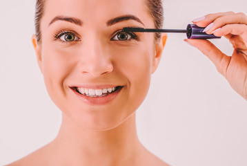 Fototapeta premium Closeup of young beautiful woman applying mascara on her long eyelashes and smiling. Happy attractive girl using mascara brush isolated on background. Putting makeup on eyes. Extension of eye lashes