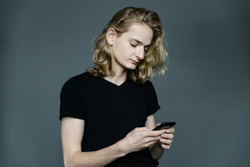 Young man with long blonde hair taking pictures of something with a phone with a strained expression on his face on a black background