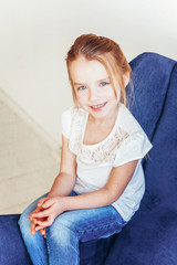 Fototapeta premium Stay Home Stay Safe. Sweet little girl in jeans and white T-shirt sitting on modern cozy blue chair relaxing in white bright living room at home indoors. Childhood schoolchildren youth relax concept.