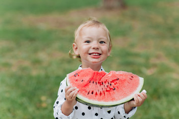 cute little girl eating big piece of watermelon on the grass in summertime. Adorable little girl playing in the garden biting a slice of watermelon