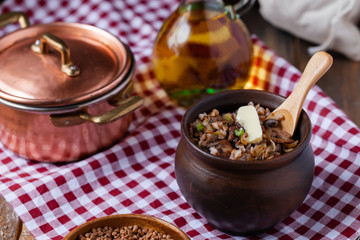 Traditional russian meal: cooked buckwheat porridge with brown butter in clay pot, served with butter, fried mushrooms, onion. Healthy food, low calories. Copper pan, wooden background, close up