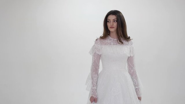young woman in white dress with lacy sleeves and bodice walks with serious face in studio slow motion close view