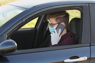 Young driver with protective surgical mask and gloves speaking by mobile phone in car, corona virus protected person