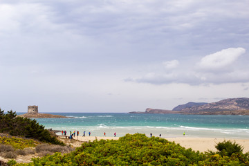 Beautifull view on the old aragonese tower and sea in La Pelosa Beach. Green trees, beach with white sand,  waves with foam and cloudly sky in Stintino, Sardinia, Italy