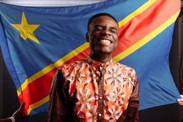 A black african man smiling and posing before the flag of the Democratic Republic of the Congo.
