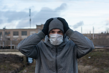 A man on the outskirts of the city against the backdrop of an abandoned industrial plant. on the face is a white rag disposable mask. He clasped his hands in pain. protection against coronavirus.