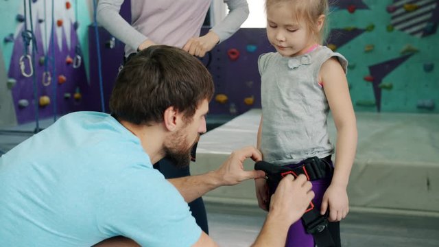 Rock-climbing instructor is helping little girl with safety harness in sports center caring for child's protection during extreme entertainment. People and hobby concept.