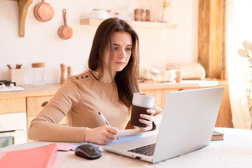 Young girl with a laptop, drinking coffee, does homework in a bright kitchen. education and business concept. remote work and freelance