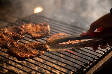 in the evening a barbecue grill on which tasty juicy steaks are grilled over an open fire, a hand holds tongs and turns the meat over.