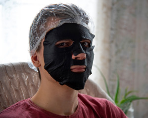 A young man in a black makeup mask and shower cap sits in a chair against a backdrop of lace curtains.