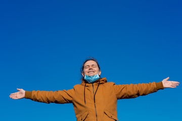 a joyful man with his hands apart, took off the mask from his face and enjoys the fresh air in freedom against a blue sky with copy space.