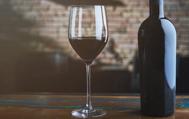 Glass of red wine and wine bottle on colorful wooden table at home, brick wall in the background.