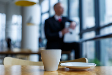 A white cup and saucer with hot aromatic coffee stands on a wooden table against the background of a sitting businessman