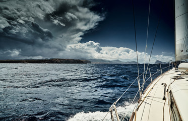 Fototapeta premium The view of the sea and mountains from the sailboat, edge of a board of the boat, slings and ropes, splashes from under the boat, sunny weather, dramatic sky