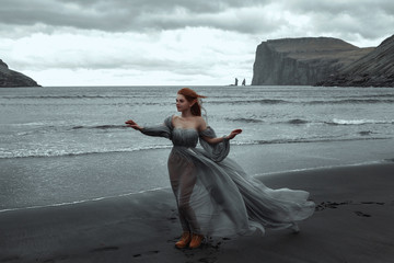 beautiful young girl with elf ears standing on the black sand in the wind near the ocean in a...