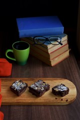 Three chocolate brownie sprinkled with castor sugar on a wooden board in the foreground with a green mug and a pile of books in the background.  A pair of glasses on the stack of books.