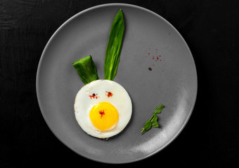 Easter bunny made of fried egg, rabbit ears made of wild garlic leaves on a ceramic plate. A healthy, fun festive breakfast. Black natural stone background, top view, space for text, minimal style