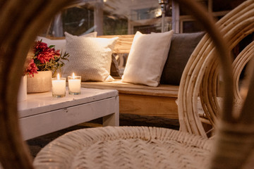 Decoracion de sala de estar con sillas de mimbre, mubles de madera, velas, flores rojas y almohadones blancos.