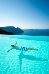 Fototapeta premium Dramatic caldera view of unrecognizable man swimming underwater in turquoise infinity pool overlooking the Mediterranean Sea in Santorini, Greece