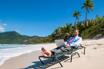 Smiling businessman working remotely on his laptop from the shore of an empty tropical beach